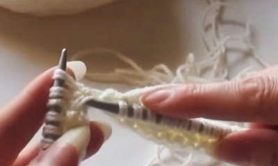 Close‑up of hands knitting with beige yarn on silver needles, showing neat rows of stitches forming a cosy fabric