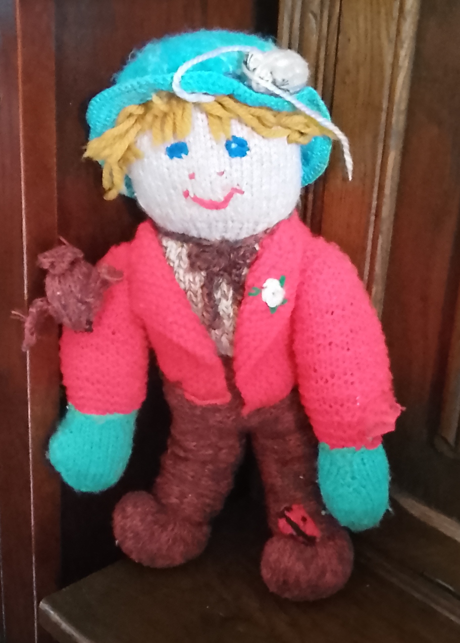 Hand‑knitted scarecrow doll with yellow hair, turquoise hat, red jacket and a small knitted bird on its arm.