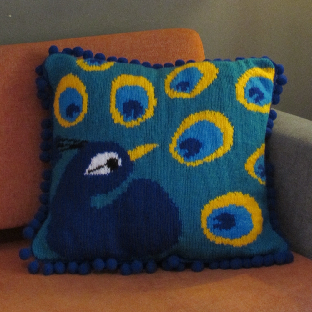 A colourful knitted peacock cushion on a sofa, illustrating the cultural meaning of knitted household items.