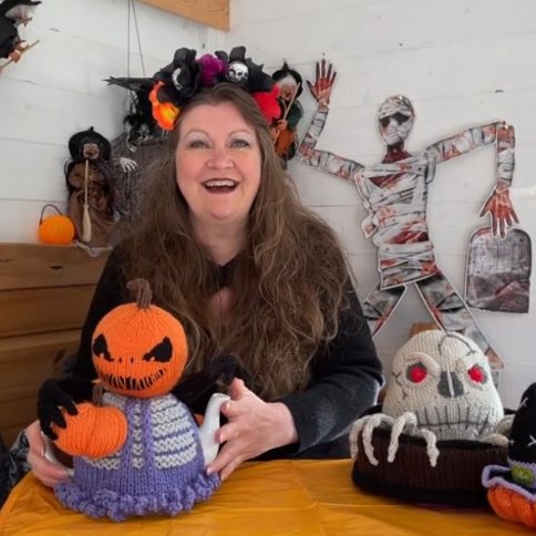 Suzy Cowper and the October TeaCosyFolk knitting vlog, surrounded by seasonal knits including Halloween-themed tea cosies and chocolate orange covers
